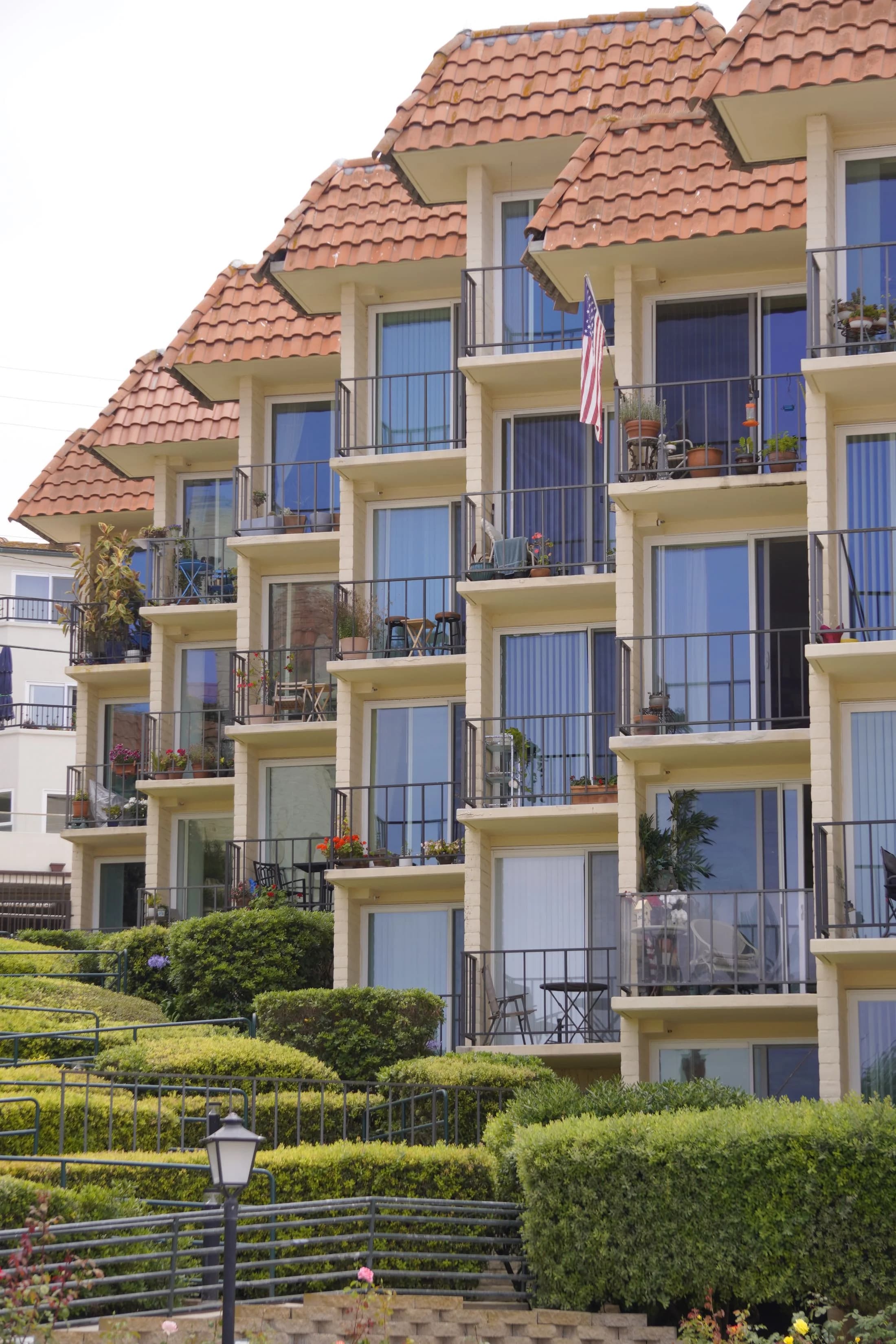 Occupied apartment building with furnished balconies, mature landscaping, and an American flag.
